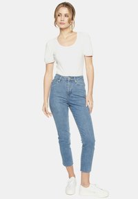 Biały krótki top z okrągłym dekoltem, połączony z niebieskimi dżinsami o wysokim stanie. Modelka nosi białe sneakersy, stojąc na jednolitym tle.