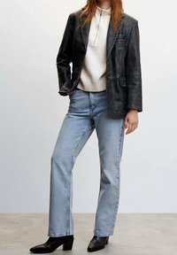 Frau in schwarzer Lederblazer, weißem Reißverschluss-Pullover, hellblauen Jeans und schwarzen Absatzstiefeln, die vor einem einfarbigen grauen Hintergrund steht.