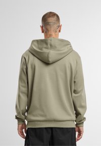 Olijfgroene hoodie met een zachte textuur, verstelbare trekkoordcapuchon en geribbelde mouwen. De achterkant toont een eenvoudig, minimalistisch ontwerp.