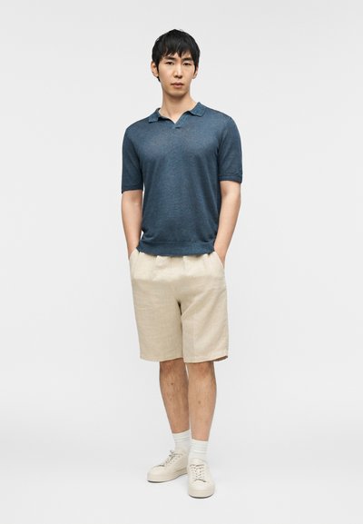 Polo i mørkeblått strikket stoff og beige linshorts. Modellen har på seg hvite sokker og beige joggesko, står med hendene i lommene.
