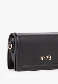 Cartera de cuero negro con textura suave, herrajes dorados y logotipo "V°73" en relieve. Presenta una forma rectangular estructurada.