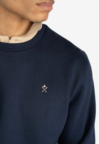 Sudadera azul marino con cuello redondo, con una camiseta de color beige claro debajo. Logo bordado en un color claro en el pecho.