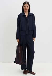 Woman with long dark hair wearing a navy jacket and pants, holding a large dark striped tote bag, standing against a white background.
