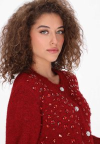 Czerwony sweter z dzianiny o teksturowej, puszystej tkaninie. Ozdobiony różnymi czerwonymi i przezroczystymi cyrkoniami, z okrągłym dekoltem i luźnym krojem.