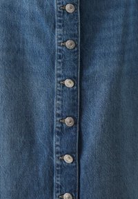 Farkkumekko, jossa on edessä napitus hopeisilla koristeilla, keskisininen pesu, kestävä rakenne ja näkyvät saumat.