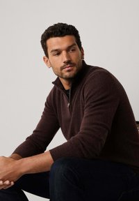 Maglione in lana marrone con zip a quarti e polsini e colletto a coste, indossato sopra una camicia blu navy. Jeans in denim scuro. Modello seduto con postura rilassata.