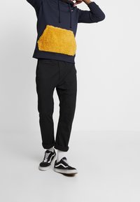 Person med marinblå hoodie med stor fluffig gul ficka, svarta uppvikta byxor, vita strumpor och svarta sneakers som står på vit golv.