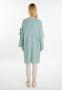 Maglione lungo a maglia di un verde chiaro, con maniche a strati a balze e spacchi laterali, abbinato a pantaloni cropped chiari e scarpe beige.