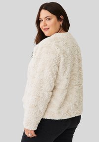 Vrouw met lang bruin haar in een crèmekleurige fluffy jas en zwarte jeans, die over haar schouder kijkt tegen een effen grijze achtergrond.