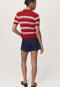 Kvinna som bär en röd- och vitrandig skjorta, mörkblå shorts och svarta ballerina, stående med ryggen mot kameran.