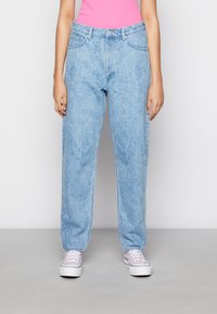 Jeans de ganga azul claro com um corte solto, apresentando uma cintura alta, passadores de cinto e bolsos frontais, acompanhados por sapatilhas cor-de-rosa.