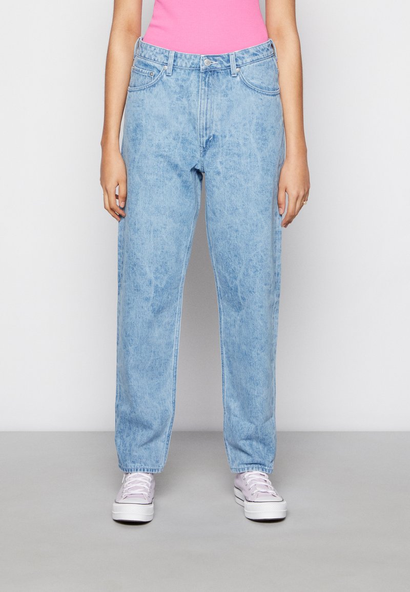 Jeans de ganga azul claro com um corte solto, apresentando uma cintura alta, passadores de cinto e bolsos frontais, acompanhados por sapatilhas cor-de-rosa.