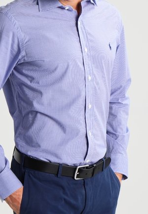 Muž nosící světle modrou kostkovanou košili zapnutou a vloženou do tmavě modrých kalhot s černým páskem, ruce v kapsách.