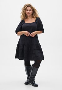 Schwarzes Kleid mit quadratischem Ausschnitt, Puffärmeln und gestuftem, rüschiertem Saum. Der Stoff wirkt weich; begleitet von kniehohen schwarzen Stiefeln.