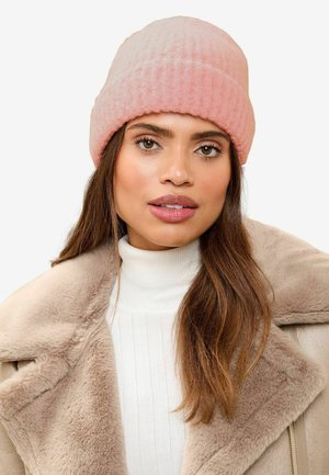Pink ribbed beanie made of soft material, featuring a folded edge. Worn with a white ribbed turtleneck and a beige shearling coat.