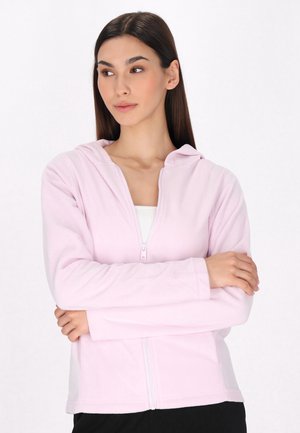 Felpa con zip di color rosa chiaro, in morbido tessuto felpato, con cappuccio e maniche lunghe, indossata sopra una maglietta bianca. Design semplice e funzionale.