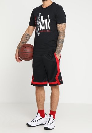 T-shirt noir Nike avec impression graphique "Dunk" en blanc et rouge. Short noir avec des accents rouges, baskets blanches et chaussettes rouges.