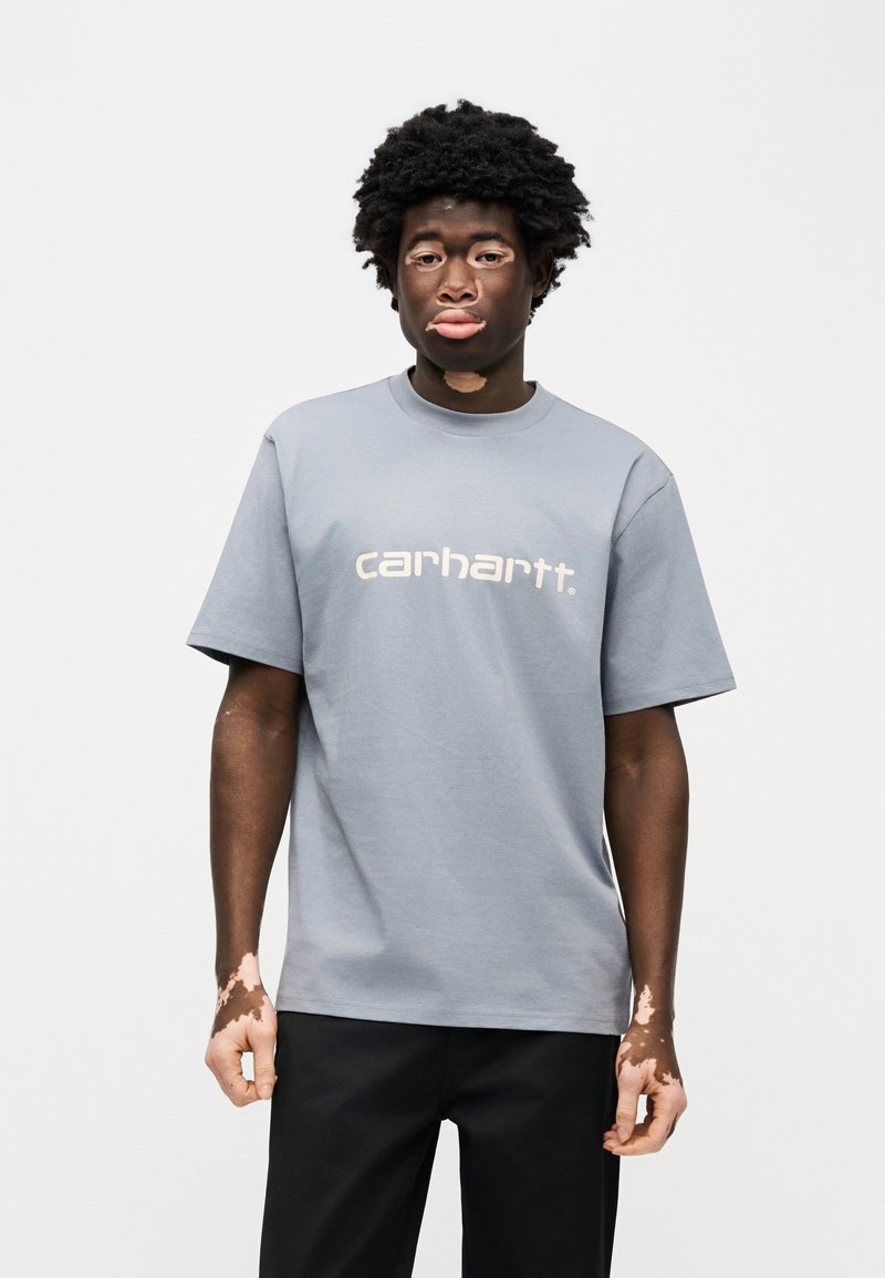 Grijs katoenen T-shirt met een ronde hals en korte mouwen. Middenvoor wit "carhartt"-logo. Draagt bij zwarte broek.