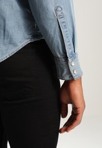 Närbild av en person som bär en ljus denimskjorta med knäppta manschetter och svarta jeans, visar bakfickan och en avslappnad hand.