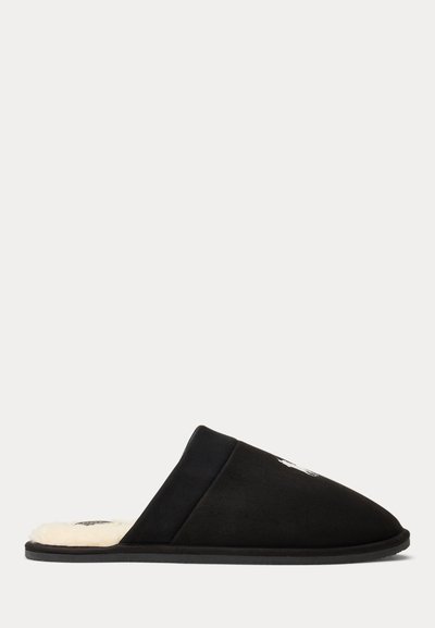 Schwarzer Slip-On-Hausschuh mit plüschigem weißem Innenfutter und flacher Sohle, von der Seite vor neutralem Hintergrund gezeigt.