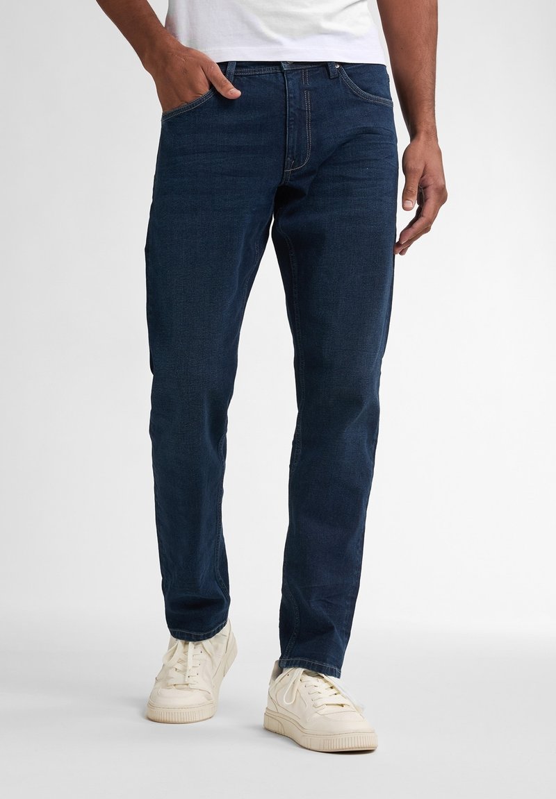 Vaqueros de mezclilla azul oscuro con corte ajustado, cinco bolsillos y detalles de costura sutil, combinados con zapatillas blancas.