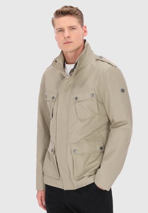 Mand iført beige jakke med fire forlommer med knapper og høj krave, poserer foran en ensfarvet lys baggrund.