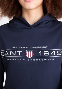 Hanorac bleumarin din amestec de bumbac, cu un grafic frontal cu "GANT 1949" și un scut, accentuat cu text alb și stele.