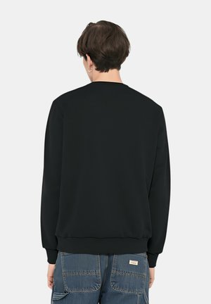 Persona che indossa una felpa nera a maniche lunghe e pantaloni cargo in denim blu, in piedi di spalle su uno sfondo chiaro e uniforme.