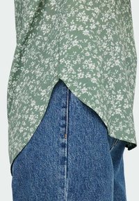 Grøn top med blomster mønster, der har en buet nederste kant og tekstureret stof; parret med blå denim jeans med en lodret søm.