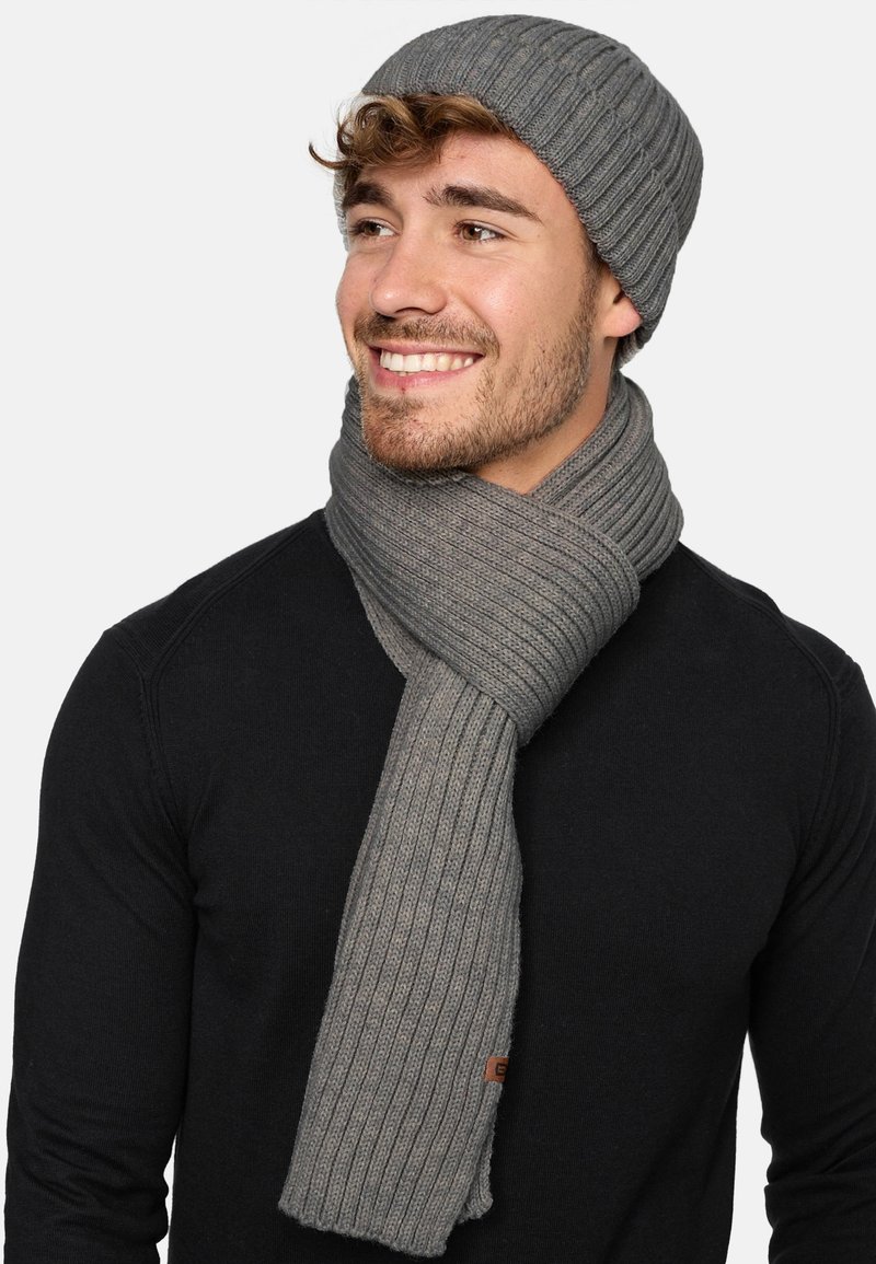 Grijze geribbelde gebreide beanie en bijpassende sjaal, met een zachte textuur. De sjaal is losjes om de nek gewikkeld, met een klein leren label.