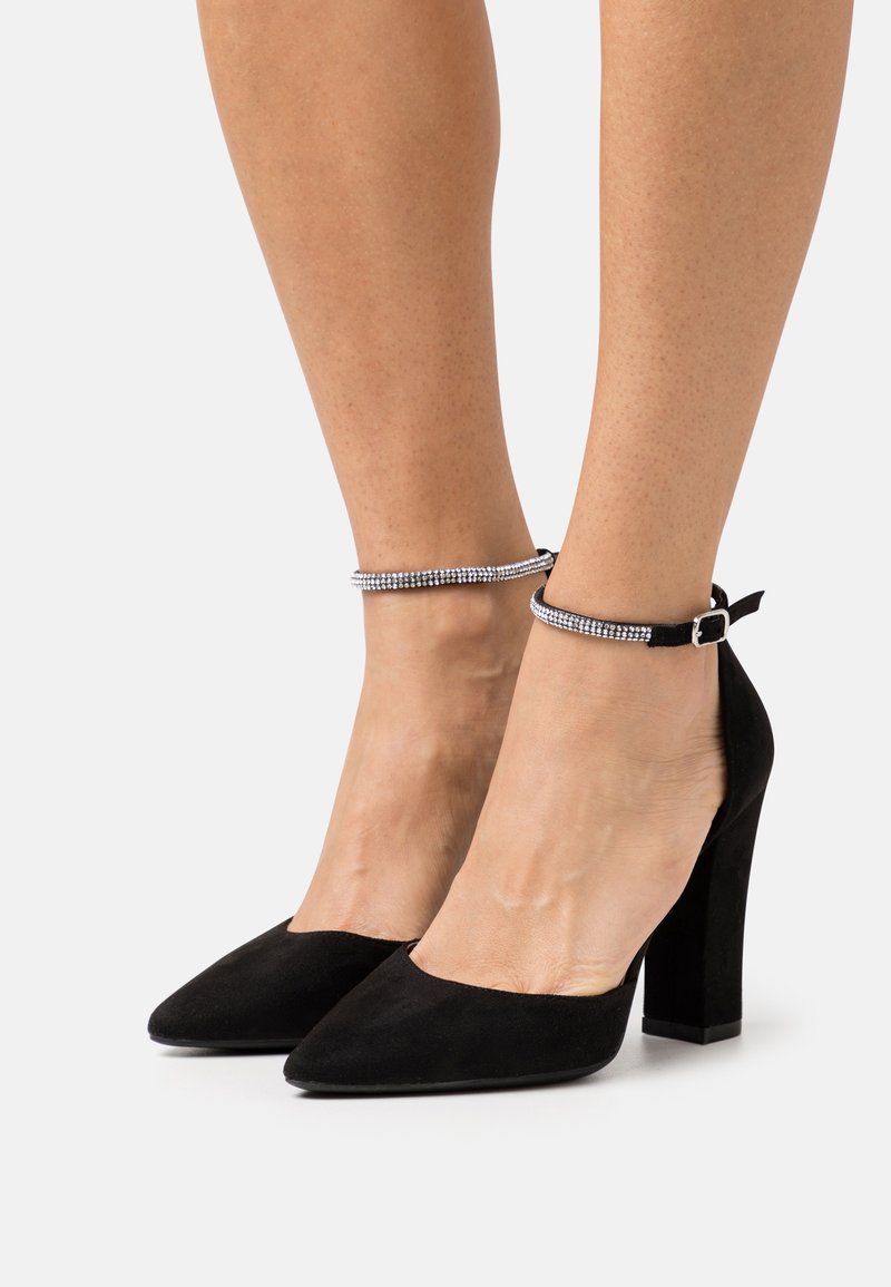 Chaussures à talons hauts en suède noir avec des orteils pointus et de fines brides de cheville ornées de petits strass sur les pieds d'une personne.