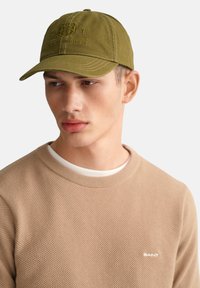 Grüne Kappe mit gesticktem Logo, gebogenem Schirm und strukturiertem Stoff. Getragen mit einem beigen, strukturierten Pullover mit Rundhalsausschnitt.