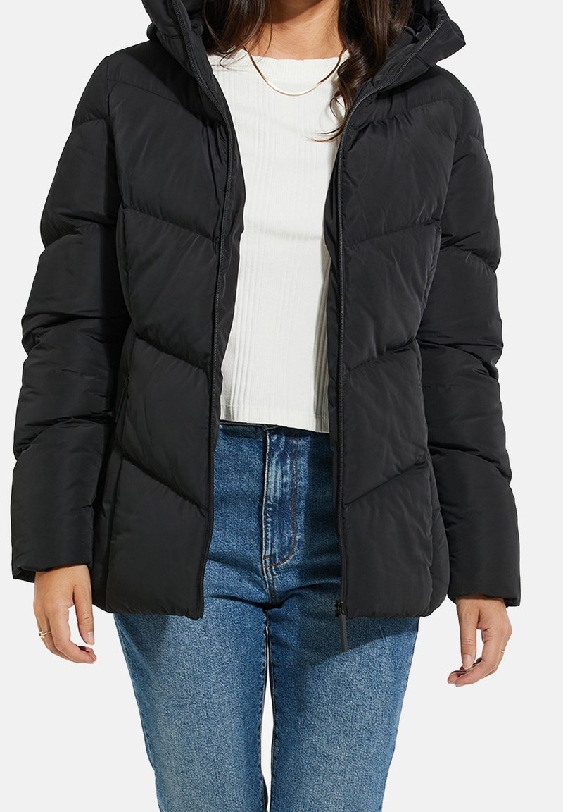 Veste matelassée noire avec capuche, design matelassé, fermeture éclair à l'avant et poignets élastiques. Portée par-dessus un haut blanc côtelé et un jean bleu.