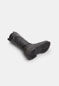 Black knee-high boot made of smooth leather with a textured rubber sole, featuring a lug pattern for traction and a pull tab at the top.