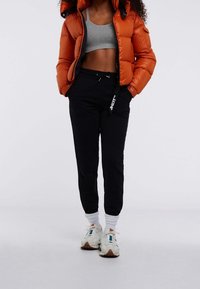 Pomarańczowa kurtka puchowa, szary krótki top, czarne spodnie dresowe ze ściągaczem, białe skarpetki i trampki z brązowymi akcentami.