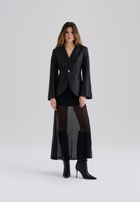 Blazer noir avec un décolleté en V profond, fermeture à boutons et manches longues, associé à une jupe plissée noire transparente et des bottines noires à talons pointus.