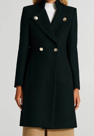 Femme portant un manteau noir à double boutonnage avec des boutons dorés sur un haut blanc et un pantalon beige.