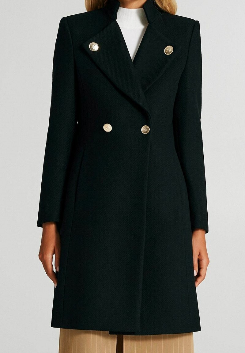 Femme portant un manteau noir à double boutonnage avec des boutons dorés sur un haut blanc et un pantalon beige.