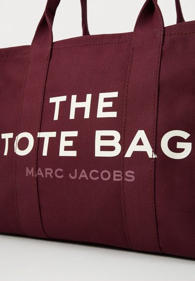 Borsa in tela bordeaux con doppio manico superiore. Caratterizzata da un testo bianco audace che legge "THE TOTE BAG" e "MARC JACOBS" in un font moderno.