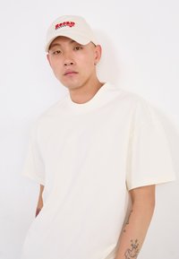 T-shirt bianca oversize con colletto rotondo abbinata a un cappellino da baseball crema con testo ricamato rosso. Design semplice e casual.