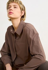 Camicia marrone con colletto e taschino frontale, realizzata in tessuto morbido. Presenta una chiusura a bottoni e una vestibilità ampia con maniche leggermente sagomate.