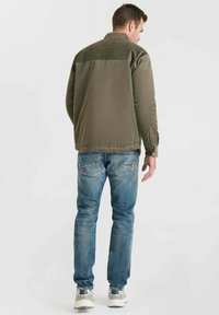 Man in olijfgroene corduroy jas, blauwe jeans en grijze sneakers, staand met de rug naar een effen achtergrond gekeerd.