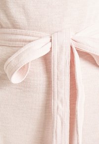 Robe rose clair avec un tissage texturé, une ceinture nouée et une finition douce. Le tissu est respirant et drape à la manière d'un vêtement ample.