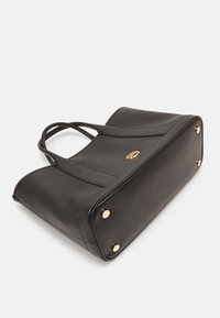 Sac tote en cuir noir avec une forme structurée, deux poignées pour l'épaule, des accents en métal doré et une surface texturée. Quatre pieds en métal à la base.
