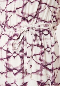 Gürtelkleid aus weißem Stoff mit lila geometrischem Muster, glatter Textur und einer Taillenschnürung mit metallicfarbenen Enden.