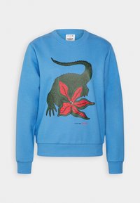 Ljusblå sweatshirt med en grafik av en grön ödla och röd blomma. Tillverkad av mjukt material med ribbade ärmslut och nederkant.