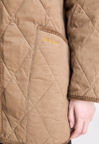Braune gesteppte Jacke mit Cordstoff-Textur, ausgestattet mit einem gelben gestickten "Barbour"-Logo auf einer Tasche und gerippten Details an den Kanten.