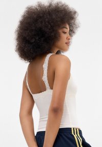 Young woman with curly hair wears a white lace-trimmed tank top and navy blue athletic pants with yellow stripes, posing sideways.