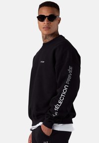 Sweat-shirt noir en tissu, avec un col rond, une coupe décontractée et un design de texte blanc sur la manche gauche et la poitrine.