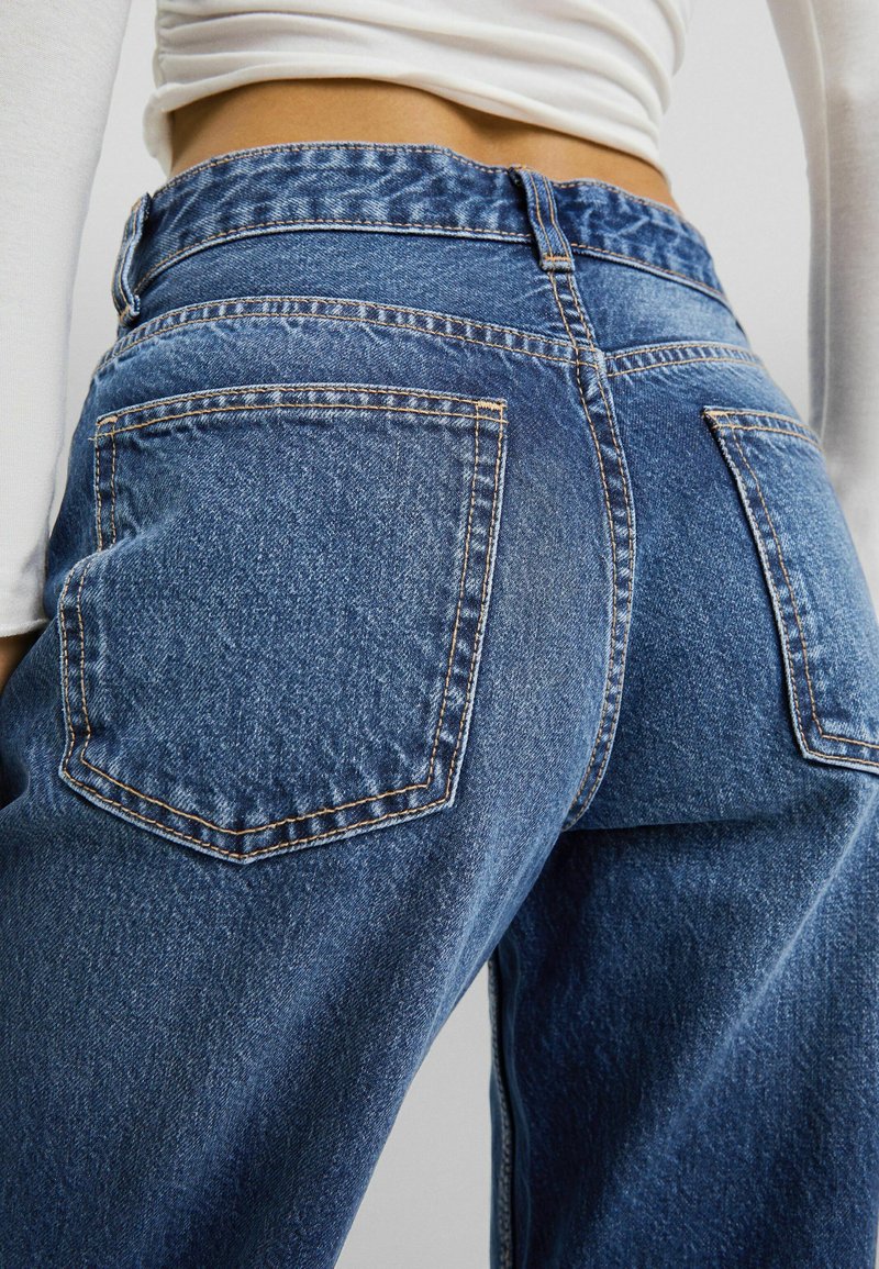 Hochgeschnittene blaue Denim-Jeans mit einer ausgewaschenen Optik, ausgestattet mit zwei Gesäßtaschen und kontrastierenden Nähten. Das Bein verjüngt sich leicht nach unten.